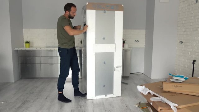 Young man unpacking new fridge from box at home
