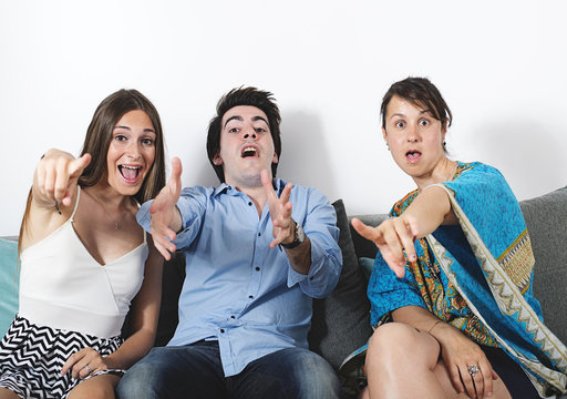 Portrait of friends, two women and one man, pointing at the camera with face and gestures of surprise.
