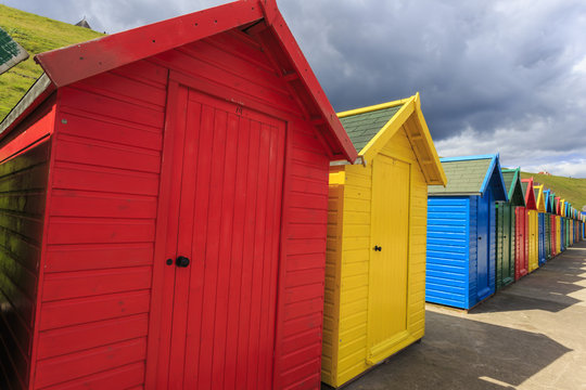Row of colourful beach huts and their shadows, with grassy cliffs, West Cliff Beach, Whitby, North Yorkshire