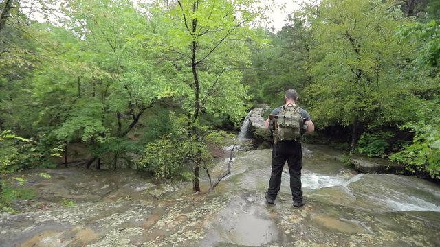A male hiker stands at a creek at the top of a waterfall, in slow motion.