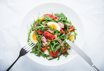 Fresh salad with tuna, tomatoes, eggs, arugula and mustard on white textured background top view. Healthy food.