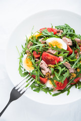 Fresh salad with tuna, tomatoes, eggs, arugula and mustard on white textured background top view. Healthy food.