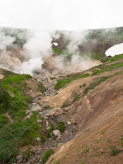 Smoking crater of the volcano Mutnovsky on Kamchatka in Russia against the background of a hill with grass and snow and sky with clouds