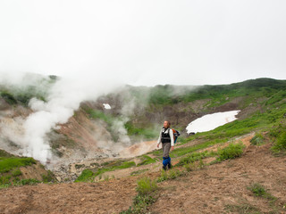 Adult woman with a backpack is walking on smoking crater of the volcano Mutnovsky on Kamchatka in Russia against the background of a hill with grass and snow and sky with clouds
