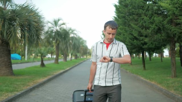 Young man tourist is with a large suitcase on wheels around the city park. Comes message on smart watches, the man stopped and dictates the answer.