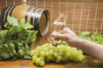 persona sujeta una copa de vino blanco, copa de vino en la ,mano, uvas sobre la mesa, barril al...