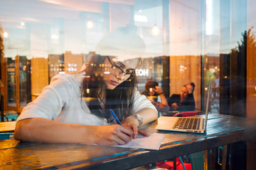 A charming student girl wearing glasses is studying in an university cafe by using a portable computer. A model look business woman is writing while sitting with a laptop behind the restaurant window.
