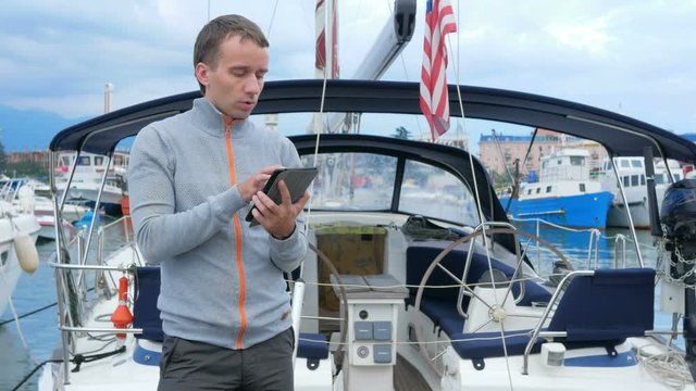 Handsome young man standing near his yacht. He checks messages on the tablet and meets friends in a social network.
