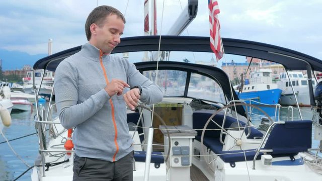 Handsome young man standing near his yacht. He checks messages on the smart watch and meets friends in a social network.