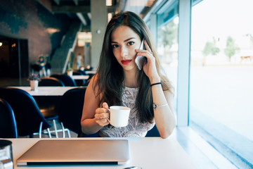 Young beautiful girl with brown hair is looking at camera while talking on a mobile phone. A charming brunette woman is sitting in a cafe with a portable computer and a smartphone.