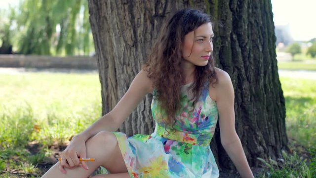 Thoughtful brunette in floral dress sitting in the park and smiling to the camera
