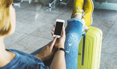 Young hipster girl sitting at airport and put feet in yellow boot on suitcase traveling, female hands using app on making smartphone in terminal area hall, tourist journey trip, mockup blank screen