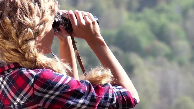 Young blonde woman tourist   on a cliff looking through binoculars on the landscape with the river. Russia, Siberia, Salair