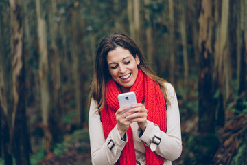 Woman texting on smartphone during a trip to the forest in winter or autumn. Brunette model wearing warm red knitted scarf and raincoat.