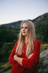 portrait of a attractive young blonde woman in red sweater with crossed arms outdoor on the background of mountain and forest