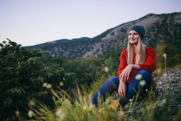 portrait of a beautiful young blonde woman in hat, red sweater, blue jeans sits outdoor on the background of mountain and forest. on the foreground blurred grass