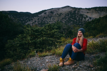 Naklejka na ściany i meble portrait of a beautiful young blonde woman in hat, red sweater, blue jeans and yellow boots sits outdoor on the background of mountain and forest