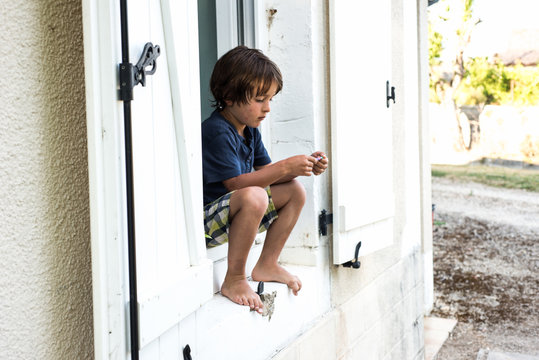 Boy sitting on holiday apartment window ledge staring at bracelet, France