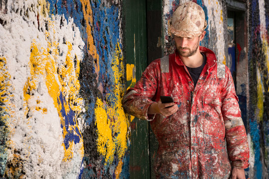 Male Ship Painter Reading Smartphone Text Leaning Against Paint Splattered Wall