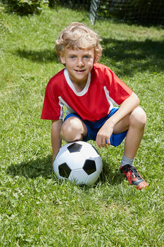 Portrait of boy wearing soccer uniform crouching with soccer ball in garden