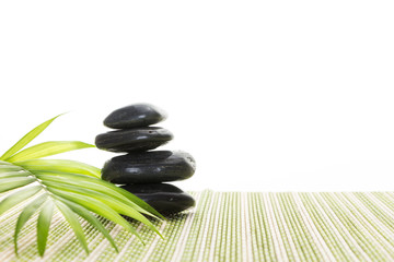Stack of black basalt balancing stones with green leaf on bamboo mat, on white background.