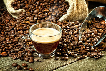  glass cup containing steaming espresso, roasted grains in jute sack on a worn wooden board background.