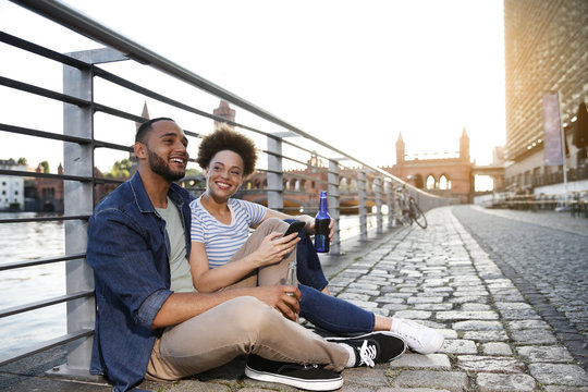 Couple sitting by river holding bottles and smart phone