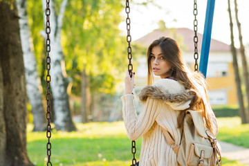 Female teenage student in sweater on a swing in park with backpack