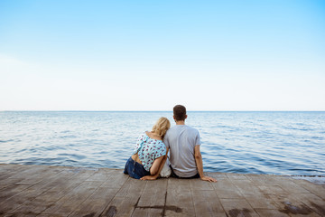 Fototapeta premium Couple sitting back to camera with French bulldog near sea.