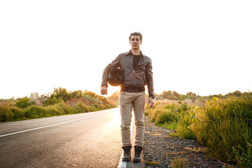 Young handsome motorbiker holding helmet, posing at countryside road. Flare sunlight background.