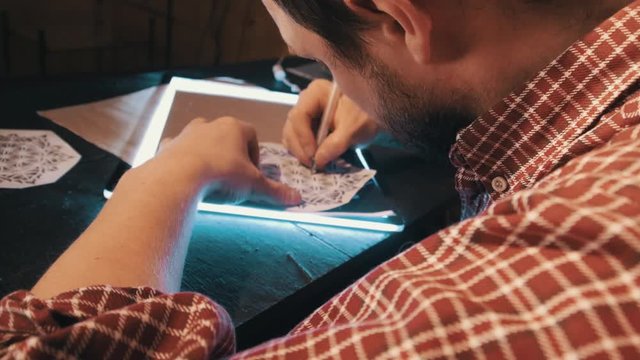 Bearded man in plaited shirt draws mandala geometrical pattern on a lightbox using copy paper