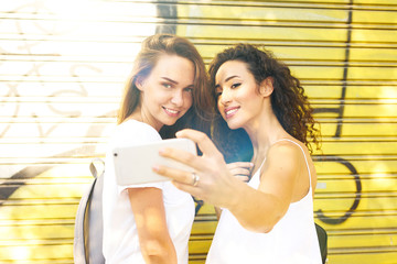 Two beautiful long haired girls wearing casual clothes are smiling at camera of a mobile phone while taking selfie beside the yellow wall with a graffiti. A waist up photo.