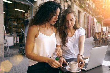 Two beautiful girls are sitting in a coffee shop with a portable computer on a summer day. Young entrepreneurs are working on a new project while drinking coffee in a cafe.