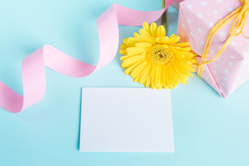 Pink dotted gift box, yellow gerbera flower and empty card over a blue background.