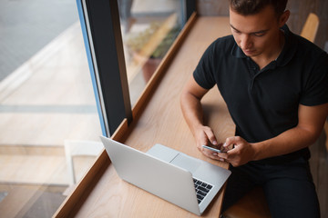 Handsome man Texting while sitting with laptop in cafe besides a window.