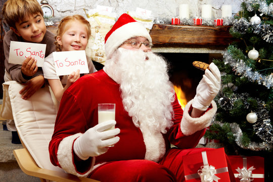 An image of Santa Claus sitting in armchair at the Christmas tree and eating cookie with milk and two children behind him