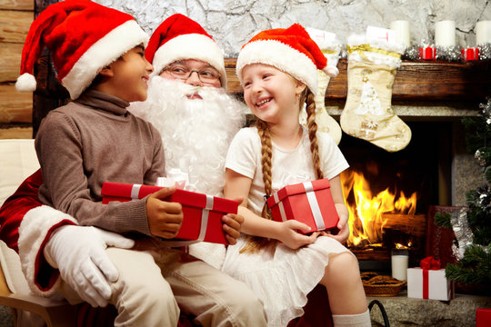 An image of Santa Claus sitting in armchair and two smiling children on his knees holding gifts