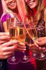 A close up of five hands clinking glasses with champagne, two girls are in the backgrounds
