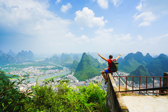 Hiker sitting on the top of hill with raised hands and enjoing view on the  valley with  city and mountains. Yangshou, China.