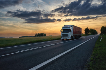 Obraz na płótnie Canvas Truck driving on an asphalt road past a cornfield under dramatic dark clouds at sunset.
