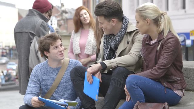 Group of students studying and talking in the city. They are wearing casual clothing and holding study materials.