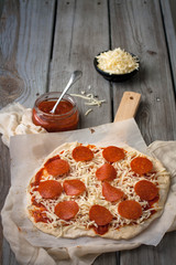 Raw pizza topped with pepperoni on a wooden pizza peel ready to going a hot oven. Tomato sauce  in the background 