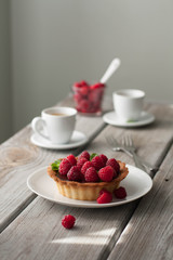 Chocolate tart with fresh raspberries and mint leaves, gray background . White ceramic teapot and cups in the background. 