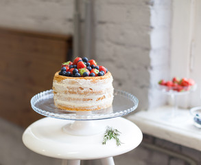 Beautiful biscuit cake with white cream decorated with strawberries and blueberries on a white pedestal stands at the window