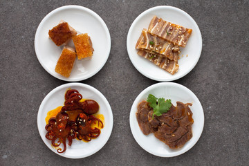 Assorted Asian appetizers on white plates:  octopus in spicy sauce, sliced beef shank, pigs ears, crispy barbecue pork . Dark gray background 