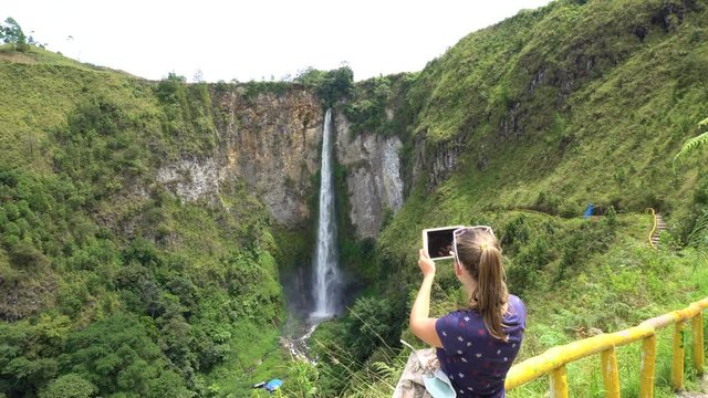 Young woman taking photo of Sipiso Piso waterfall, North Sumatra, Indonesia. Female traveler living active lifestyle