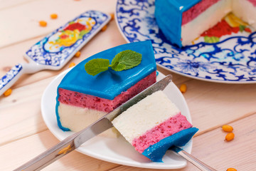 Piece of cake decorated with blue mirror glaze, mint and sea ​​buckthorn on wooden background. Homemade bright mousse cake.