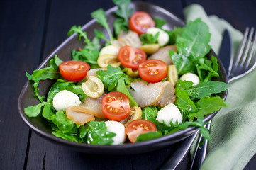 Tasty salad with slices of the dried beef, a mozzarella, olives, cherry tomatoes and arugula with olive oil and spices. Food composition on a dark wooden background. Mediterranean kitchen