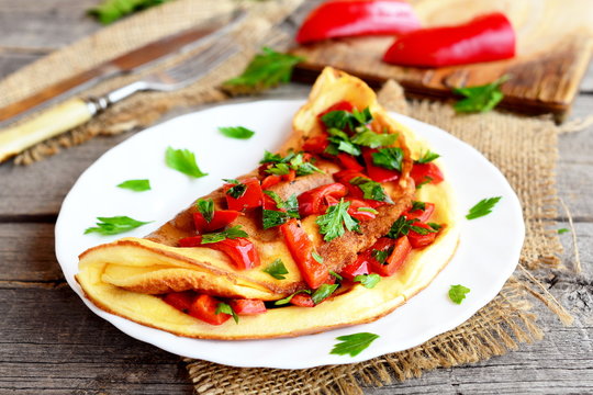 Omelette with vegetables on a plate, fork and knife on old wooden background. Colorful omelette stuffed with roasted red pepper and chopped fresh parsley. Breakfast egg recipe. Closeup