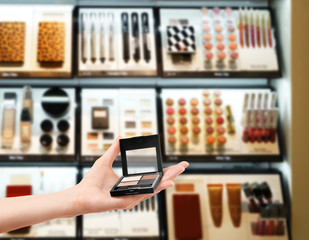 Female hand holding eyeshadow palette on blurred cosmetics shelf background. Beauty concept.